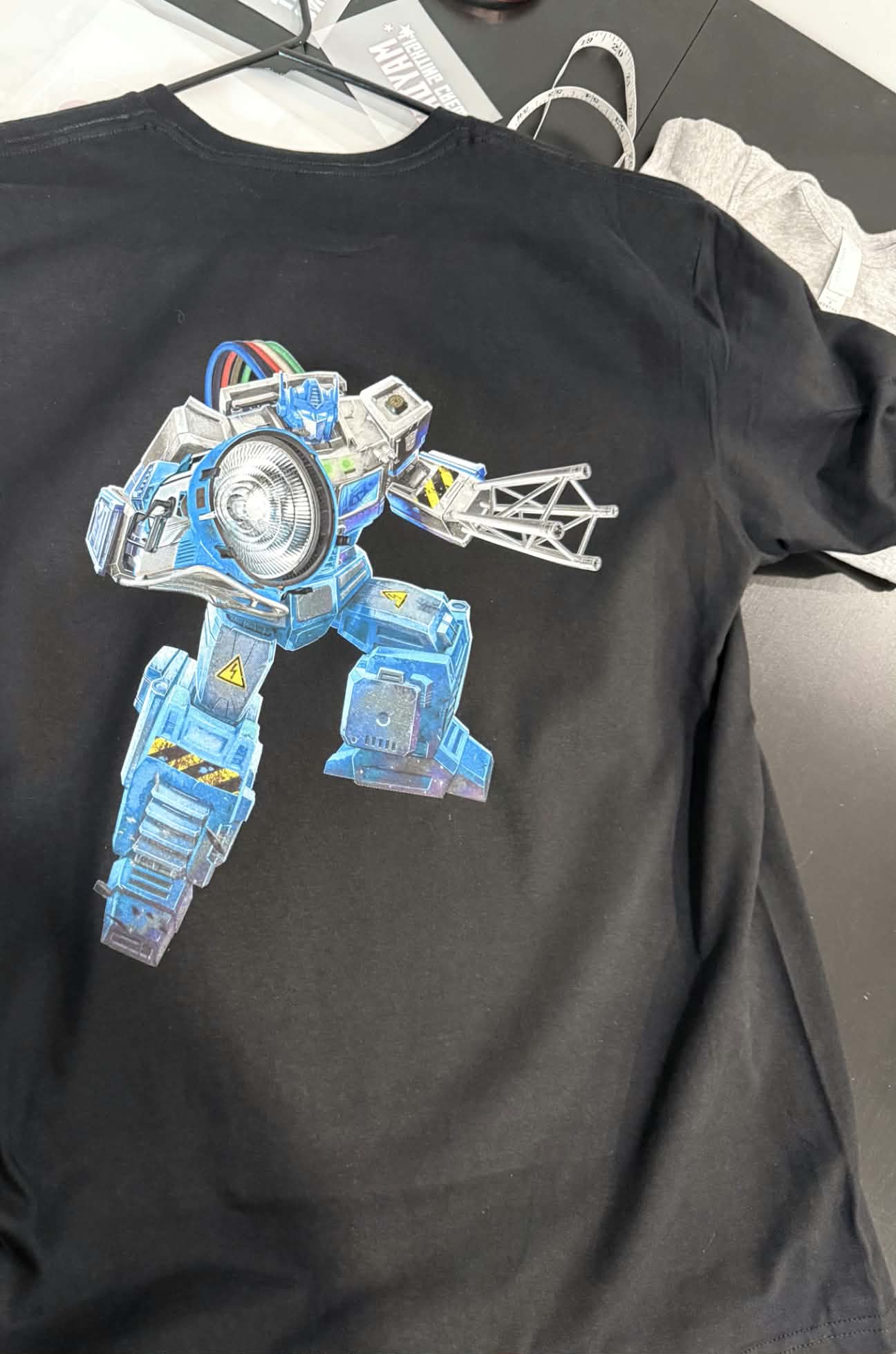 Un t-shirt noir imprimé d'un robot bleu et argent très détaillé tenant une section de truss d'éclairage — couleurs pleines et opaques sur le tissu foncé.